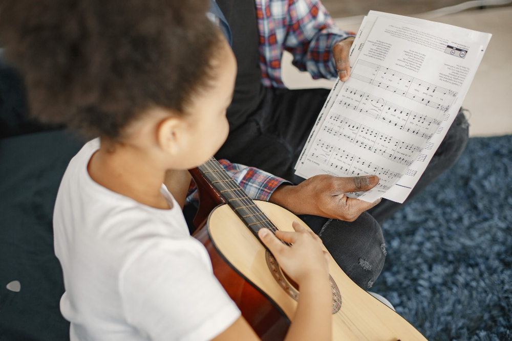 Criança aprendendo a tocar violão com acompanhamento de um professor e partitura durante aula de música.